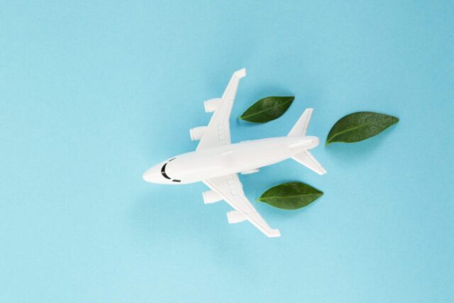 White airplane model emitting fresh green leaves on blue background. Sustainable travel; clean and green energy; and biofuel for aviation industry concept.
